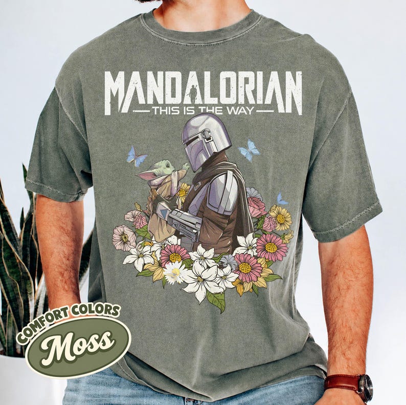 May include: Moss green t-shirt featuring a graphic of The Mandalorian and Grogu surrounded by flowers and butterflies. The text "MANDALORIAN - THIS IS THE WAY" is printed above the graphic. The shirt has a Comfort Colors label.