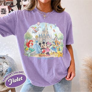 Op de afbeelding: Een violetkleurig T-shirt met korte mouwen en een grafische print van een kasteel en diverse tekenfilmfiguren, waaronder een zeemeermin, een muis en een eend. De tekst Magic Kingdom staat in het lichtblauw onder de figuren.