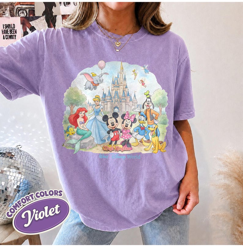 Op de afbeelding: Een violetkleurig T-shirt met korte mouwen en een grafische print van een kasteel en diverse tekenfilmfiguren, waaronder een zeemeermin, een muis en een eend. De tekst Magic Kingdom staat in het lichtblauw onder de figuren.