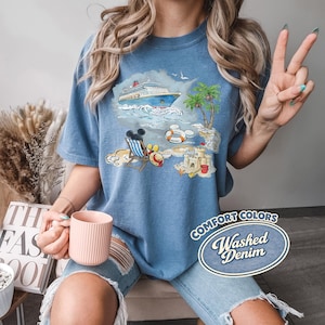 Op de afbeelding: Blauw Comfort Colors T-shirt met een strandsc&egrave;ne-afbeelding met een cruiseschip, palmbomen en een zandkasteel. De afbeelding bevat Mickey Mouse en de tekst "Washed Denim".