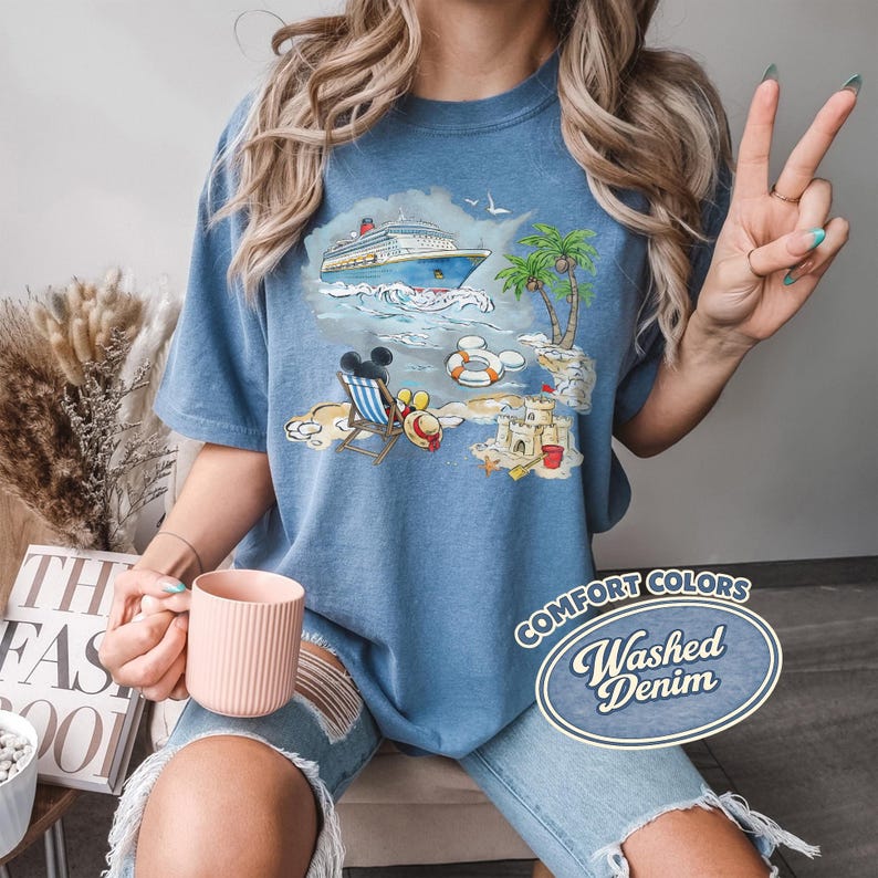Op de afbeelding: Blauw Comfort Colors T-shirt met een strandsc&egrave;ne-afbeelding met een cruiseschip, palmbomen en een zandkasteel. De afbeelding bevat Mickey Mouse en de tekst "Washed Denim".