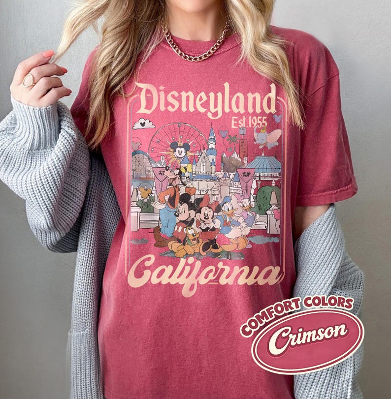 Może przedstawiać: Karmelowa koszulka z grafiką Disneyland z Myszką Miki, Minnie i Kaczorem Donaldem. Projekt zawiera napisy "Disneyland California Est. 1955" w stylu vintage. Koszulka jest częścią kolekcji Comfort Colors.