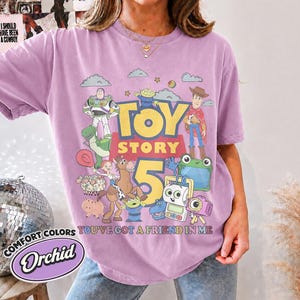 May include: A light purple t-shirt featuring a graphic print of Toy Story characters including Buzz Lightyear, Woody, Jessie, Rex, and others. The text reads Toy Story 5 and You've Got A Friend In Me. The shirt has a crew neckline and short sleeves.