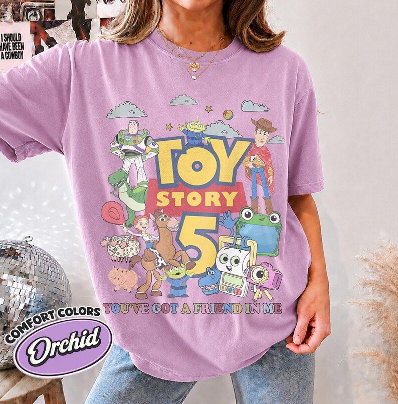 May include: A light purple t-shirt featuring a graphic print of Toy Story characters including Buzz Lightyear, Woody, Jessie, Rex, and others. The text reads Toy Story 5 and You've Got A Friend In Me. The shirt has a crew neckline and short sleeves.