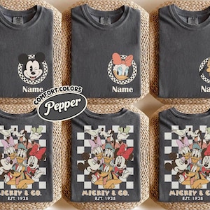 May include: Dark gray t-shirts featuring Disney characters Mickey Mouse, Daisy Duck, and Pluto. The shirts have a round neck and short sleeves. Some shirts have a character's face in a checkered circle, with the word "Name" below. Other shirts have a group of characters with the text "MICKEY & CO. EST. 1928".