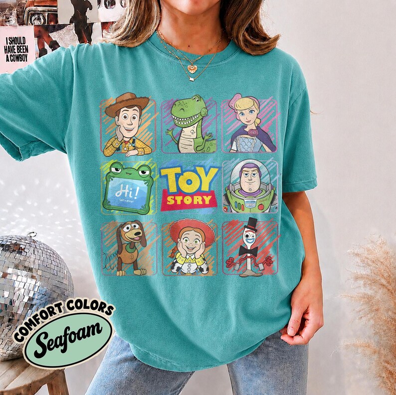 May include: A seafoam green short-sleeve t-shirt featuring a grid of Toy Story characters including Woody, Rex, Bo Peep, a green frog, Buzz Lightyear, Slinky Dog, Jessie, and Forky, with the text Toy Story in the center of the graphic design.