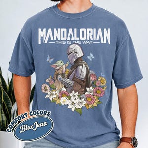 May include: Blue t-shirt with a graphic of The Mandalorian and Grogu surrounded by flowers and butterflies. The text "MANDALORIAN - THIS IS THE WAY" is printed above the image. The shirt has a "Comfort Colors Blue Jean" label.