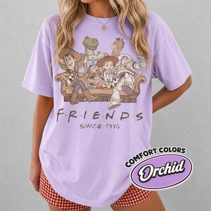 Puede incluir: Una camiseta lila con un gr&aacute;fico de personajes de Toy Story sentados en un sof&aacute;. El texto "FRIENDS SINCE 1995" est&aacute; impreso debajo del gr&aacute;fico. La camiseta tiene una etiqueta Comfort Colors Orchid.