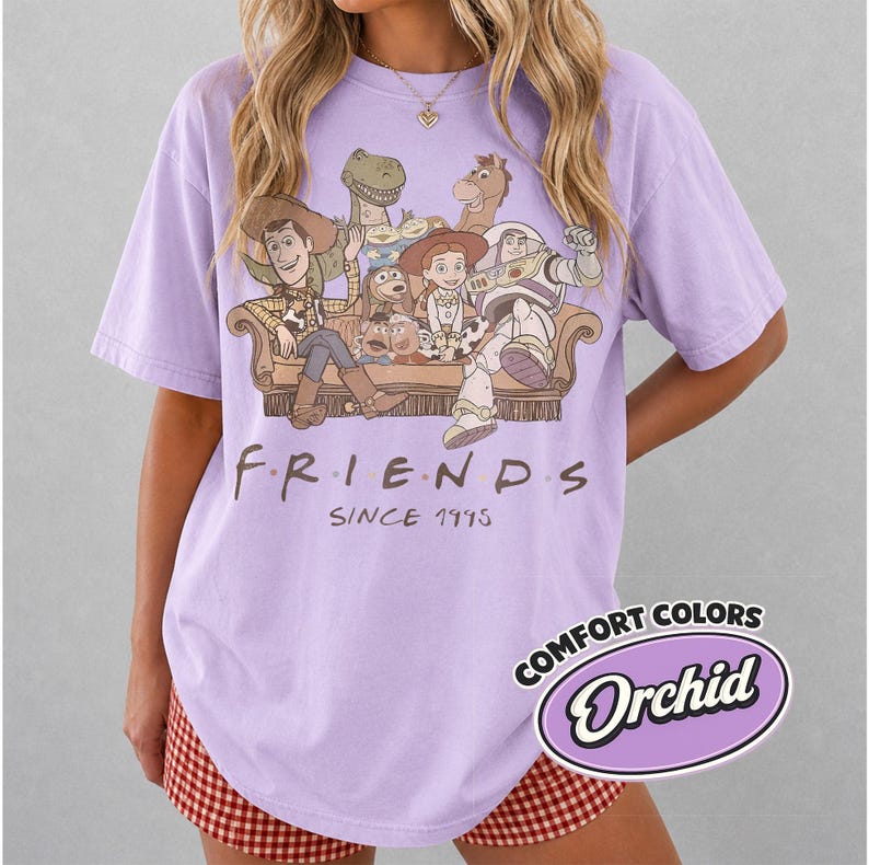Puede incluir: Una camiseta lila con un gr&aacute;fico de personajes de Toy Story sentados en un sof&aacute;. El texto "FRIENDS SINCE 1995" est&aacute; impreso debajo del gr&aacute;fico. La camiseta tiene una etiqueta Comfort Colors Orchid.