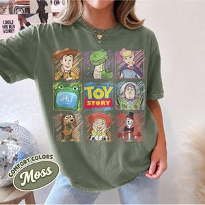 May include: A moss green t-shirt featuring a grid of Toy Story characters including Woody, Rex, Bo Peep, a green alien, Buzz Lightyear, Slinky Dog, Jessie, and Forky, with the Toy Story logo in the center and a small green tablet displaying Hi.