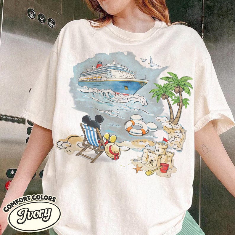 Op de afbeelding: Ivoor kleurig T-shirt met een kleurrijke strandsc&egrave;ne met een cruiseschip, palmbomen en Mickey Mouse. Het shirt heeft de woorden "Comfort Colors" en "Ivory" erop gedrukt.