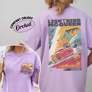 May include: A light purple t-shirt featuring a graphic of Lightning McQueen from the Cars movie. The design includes the number 95, the text "Lightning McQueen Racing Series", and a racing scene. The shirt also has the text "Comfort Colors Orchid".