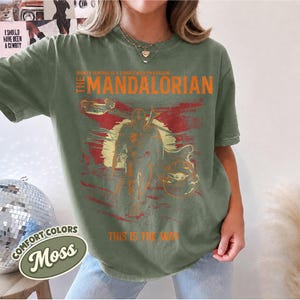May include: An olive green short-sleeve t-shirt featuring a graphic print of The Mandalorian. The design includes the text "Bounty hunting is a complicated profession," "The Mandalorian," and "This is the way" in orange, with a sunset background.