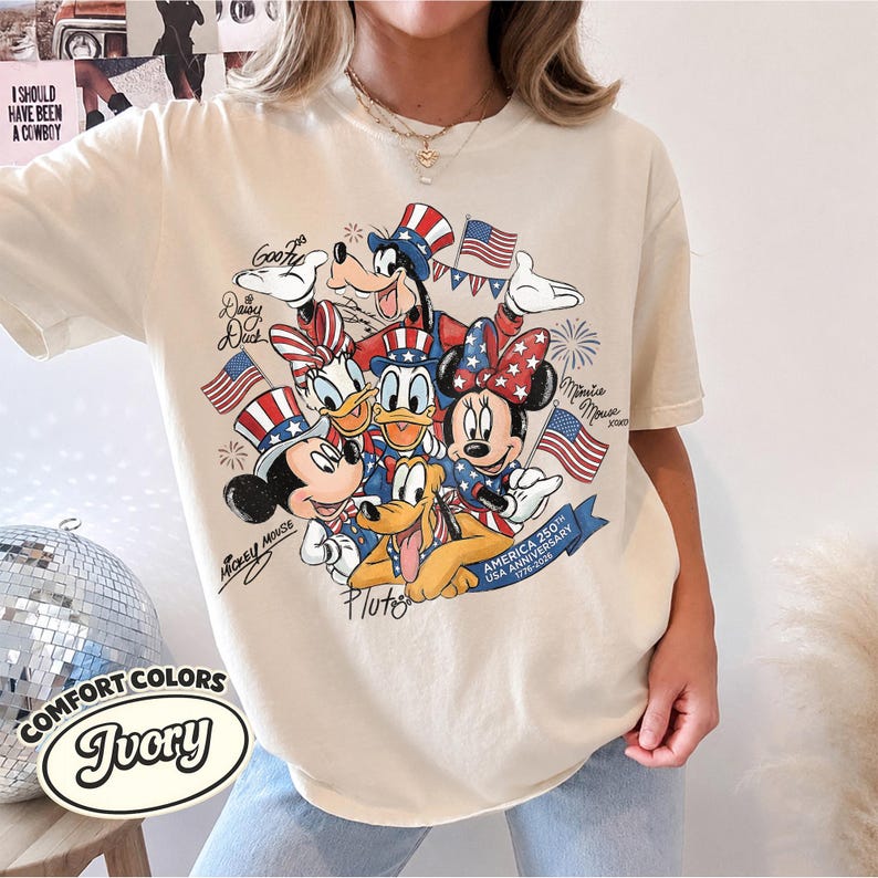 May include: Ivory-colored t-shirt featuring a graphic of Disney characters Mickey Mouse, Minnie Mouse, Donald Duck, Daisy Duck, Goofy, and Pluto, celebrating America's 250th anniversary. The design includes American flags and patriotic details.