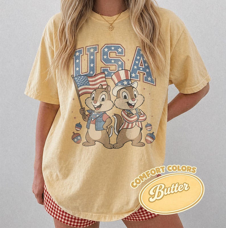 May include: Yellow t-shirt with a graphic of two cartoon chipmunks in patriotic attire, the word "USA" in blue letters with stars, and an American flag. The shirt also has the words "Comfort Colors Butter" in a decorative oval.