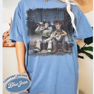 May include: Blue jean Comfort Colors t-shirt featuring a distressed graphic of Buzz Lightyear and Woody from Toy Story. The characters are depicted sitting together. The shirt has a relaxed fit and short sleeves. A logo that says "Comfort Colors Blue Jean" is on the bottom left.