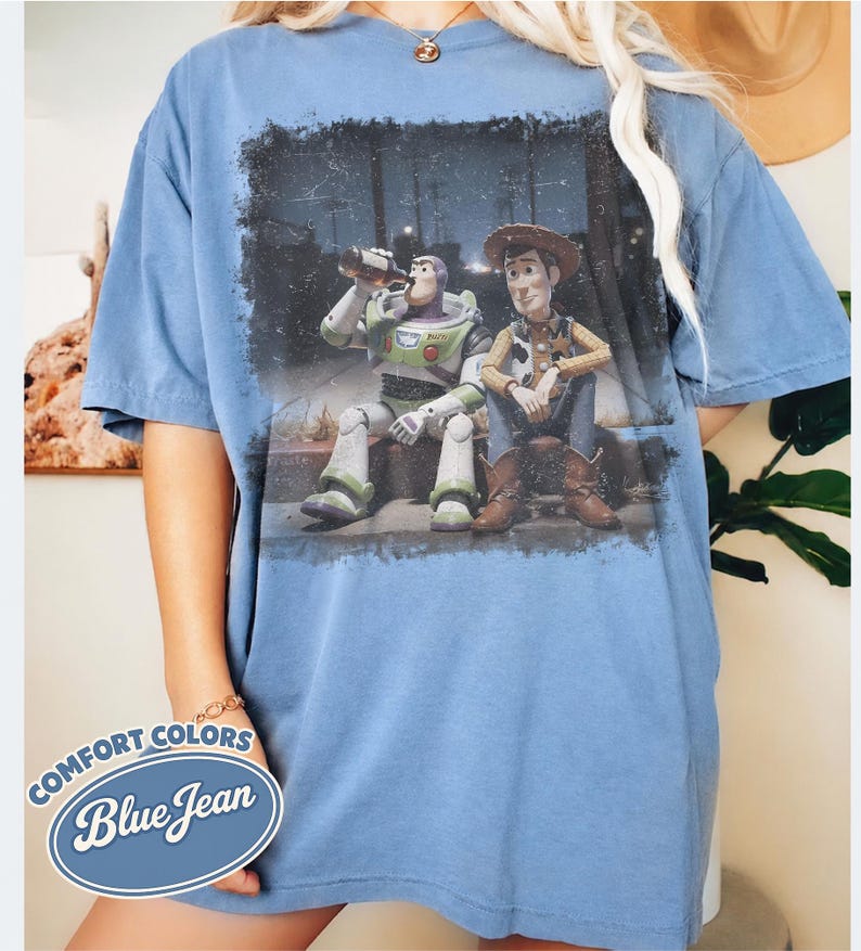May include: Blue jean Comfort Colors t-shirt featuring a distressed graphic of Buzz Lightyear and Woody from Toy Story. The characters are depicted sitting together. The shirt has a relaxed fit and short sleeves. A logo that says "Comfort Colors Blue Jean" is on the bottom left.