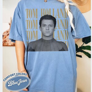 Peut inclure: T-shirt bleu clair &agrave; manches courtes avec le nom "TOM HOLLAND" imprim&eacute; en or verticalement trois fois. Une image en noir et blanc de Tom Holland en costume de Spider-Man est centr&eacute;e sur le t-shirt. Les mots "COMFORT COLORS" et "Blue Jean" sont imprim&eacute;s sur un ovale bleu.