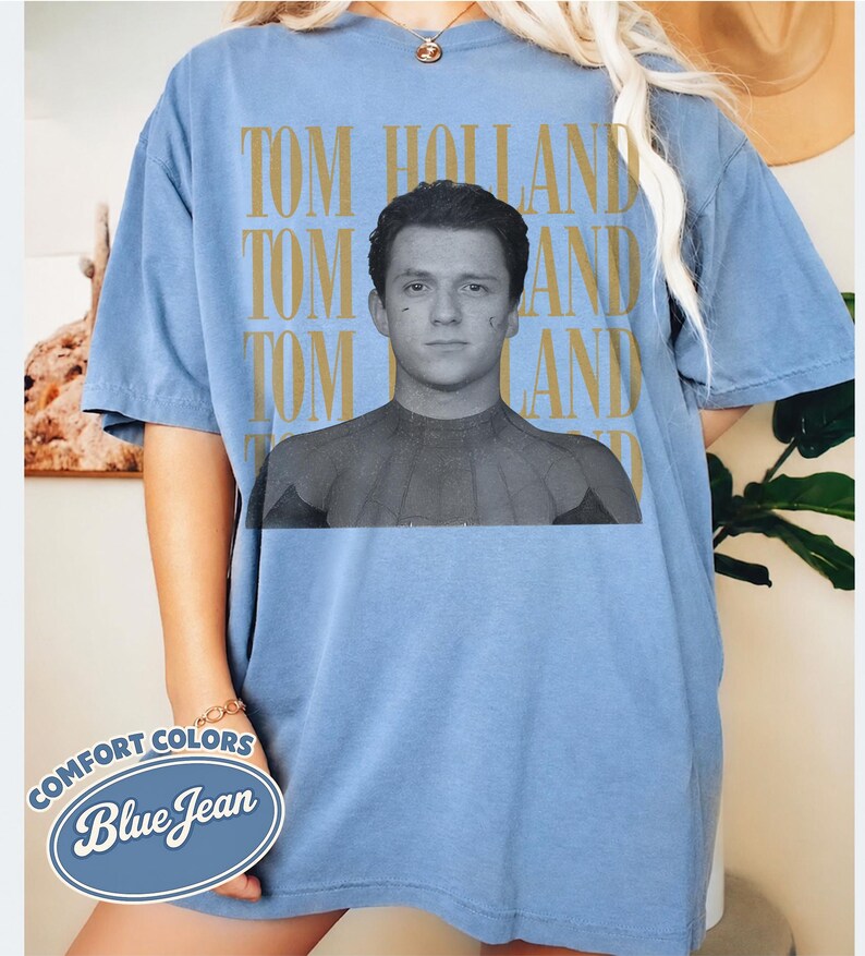 Peut inclure: T-shirt bleu clair &agrave; manches courtes avec le nom "TOM HOLLAND" imprim&eacute; en or verticalement trois fois. Une image en noir et blanc de Tom Holland en costume de Spider-Man est centr&eacute;e sur le t-shirt. Les mots "COMFORT COLORS" et "Blue Jean" sont imprim&eacute;s sur un ovale bleu.