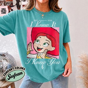 May include: A teal short-sleeve t-shirt featuring a graphic of a cartoon character with red hair and a red hat. The text reads EST 2026 I Knew It I Knew You with a musical note symbol. The shirt has a crew neckline and a relaxed fit design.