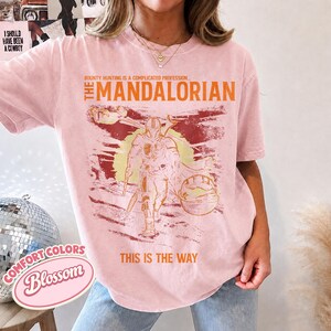 May include: A light pink short-sleeve t-shirt featuring a graphic print of The Mandalorian. The text reads, "Bounty hunting is a complicated profession... The Mandalorian" and "This is the way." The design includes a sun, a ship, and a character.