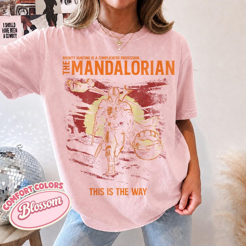 May include: A light pink short-sleeve t-shirt featuring a graphic print of The Mandalorian. The text reads, "Bounty hunting is a complicated profession... The Mandalorian" and "This is the way." The design includes a sun, a ship, and a character.