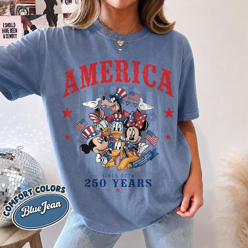 May include: A blue jean colored t-shirt featuring a graphic of Disney characters including Mickey, Minnie, Donald, Daisy, Goofy, and Pluto wearing patriotic hats. The text reads AMERICA, SINCE 1776, 250 YEARS, and AMERICA 250TH USA ANNIVERSARY.