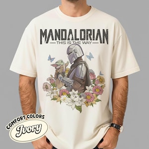 May include: Ivory-colored t-shirt featuring a graphic of The Mandalorian and Grogu surrounded by flowers and butterflies. The text "MANDALORIAN - THIS IS THE WAY" is printed across the top. The shirt has a Comfort Colors label.