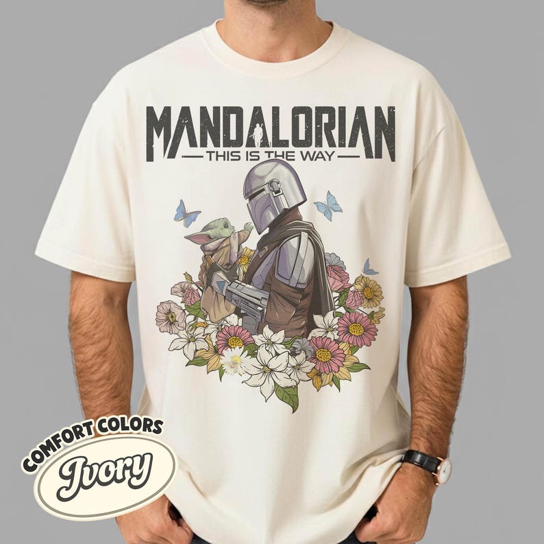 May include: Ivory-colored t-shirt featuring a graphic of The Mandalorian and Grogu surrounded by flowers and butterflies. The text "MANDALORIAN - THIS IS THE WAY" is printed across the top. The shirt has a Comfort Colors label.