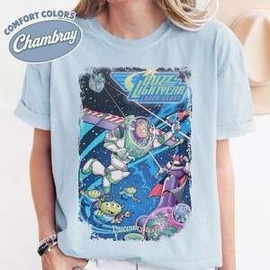 Pode incluir: Uma t-shirt azul-clara com uma estampa gr&aacute;fica de Buzz Lightyear, Zurg e alien&iacute;genas verdes no espa&ccedil;o. O design inclui o texto Buzz Lightyear Laser Blast e Discoveryland. A t-shirt tem uma etiqueta Comfort Colors Chambray.