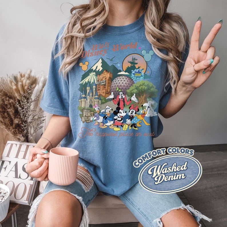 Pu&ograve; includere: T-shirt blu Comfort Colors con una grafica colorata di personaggi e monumenti di Disney World. La maglietta include il testo "Walt Disney World" e "The happiest place on earth". La maglietta &egrave; etichettata come "Washed Denim".