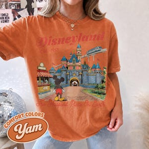 May include: An orange short-sleeve t-shirt featuring a graphic print of a castle, a monorail, and a cartoon mouse. The text reads Disneyland Est. 1955 and The Happiest Place On Earth. The shirt has a crew neckline and a relaxed, vintage-style fit.