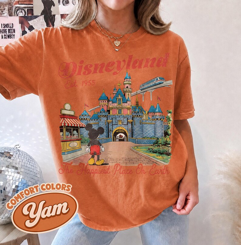 May include: An orange short-sleeve t-shirt featuring a graphic print of a castle, a monorail, and a cartoon mouse. The text reads Disneyland Est. 1955 and The Happiest Place On Earth. The shirt has a crew neckline and a relaxed, vintage-style fit.