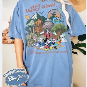 Pu&ograve; includere: T-shirt Comfort Colors blu jeans con grafica colorata di personaggi e monumenti di Disney World. La maglietta include il testo "Walt Disney World" e "The happiest place on earth."