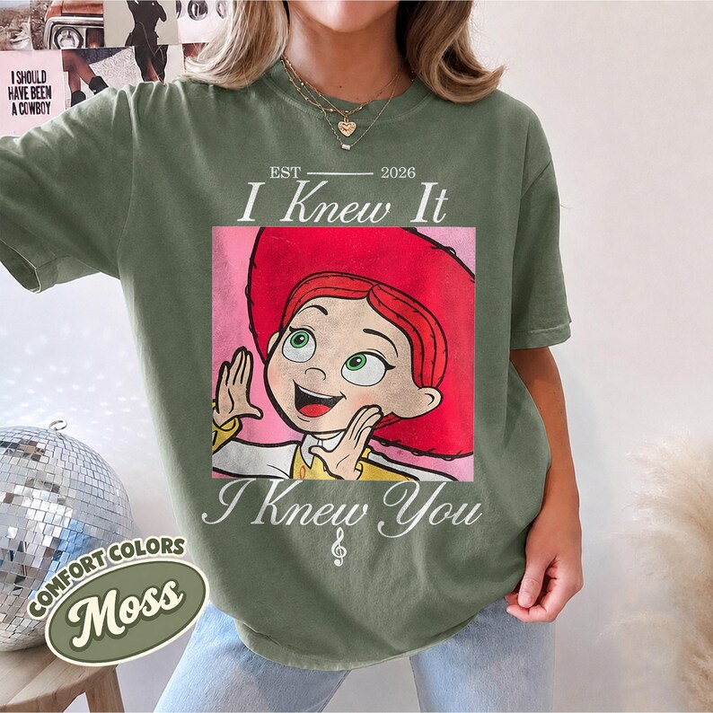 May include: A moss green short-sleeve t-shirt featuring a graphic of a red-haired character in a red hat. The text reads EST 2026 I Knew It I Knew You with a musical note symbol. The design has a distressed, vintage-inspired aesthetic.