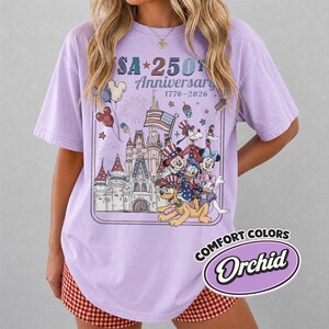 Puede incluir: Una camiseta de color lila con un gr&aacute;fico que celebra el 250 aniversario de EE. UU. El dise&ntilde;o presenta un castillo, fuegos artificiales, la bandera estadounidense y personajes de dibujos animados. La camiseta est&aacute; etiquetada como "Comfort Colors" y "Orchid".