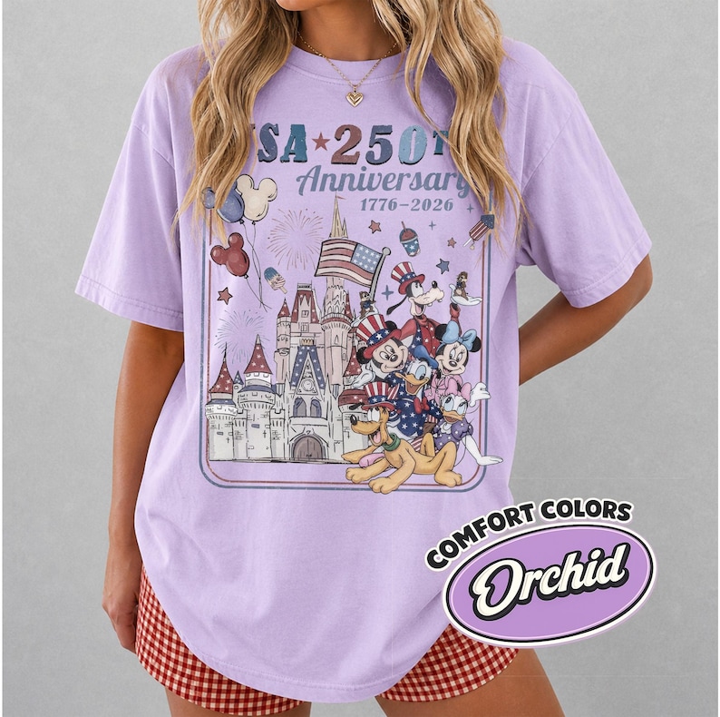 Puede incluir: Una camiseta de color lila con un gr&aacute;fico que celebra el 250 aniversario de EE. UU. El dise&ntilde;o presenta un castillo, fuegos artificiales, la bandera estadounidense y personajes de dibujos animados. La camiseta est&aacute; etiquetada como "Comfort Colors" y "Orchid".