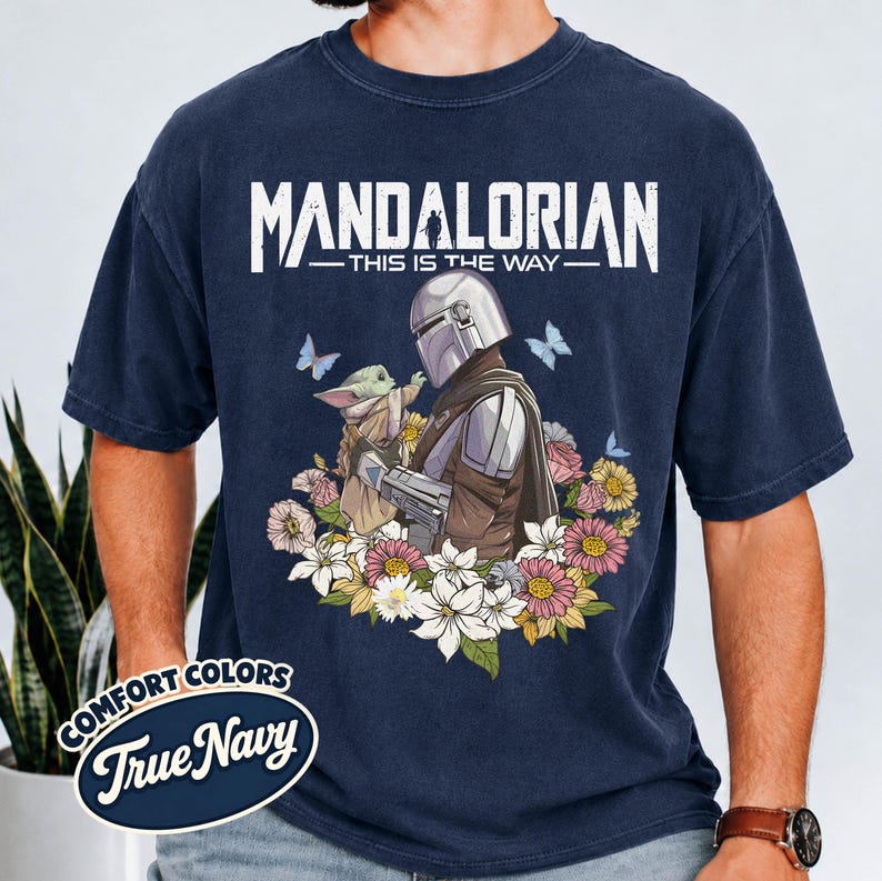 May include: Navy blue t-shirt featuring a graphic of The Mandalorian and Grogu surrounded by flowers and butterflies. The text "MANDALORIAN - THIS IS THE WAY" is printed across the top. The shirt is a Comfort Colors True Navy.