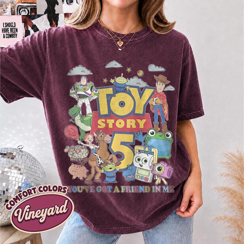 May include: A maroon short-sleeve t-shirt featuring a graphic print of Toy Story characters including Buzz Lightyear, Woody, and Rex. The design includes the text "TOY STORY 5" and "YOU'VE GOT A FRIEND IN ME" in yellow and blue lettering.