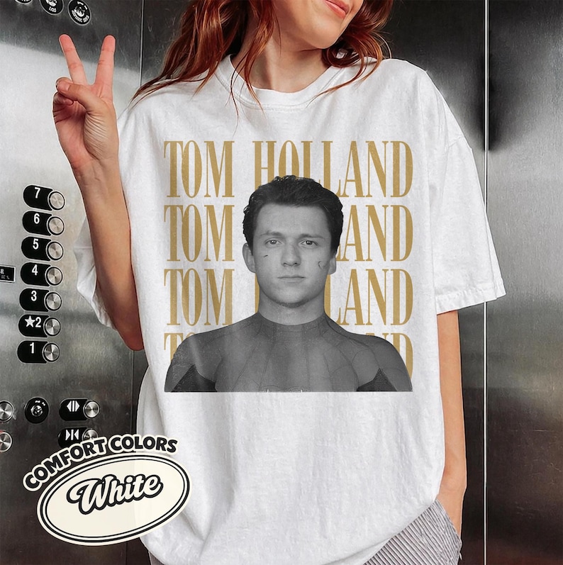 Peut inclure: T-shirt blanc Comfort Colors avec une image en noir et blanc de Tom Holland et le texte dor&eacute; "TOM HOLLAND". Le t-shirt porte &eacute;galement le logo Comfort Colors dans un ovale blanc.