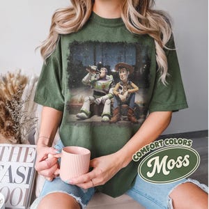 May include: Moss green t-shirt featuring a graphic of Buzz Lightyear and Woody from Toy Story. The characters are depicted sitting together. The shirt has a distressed look and the words "Comfort Colors" and "Moss" are printed on the shirt.