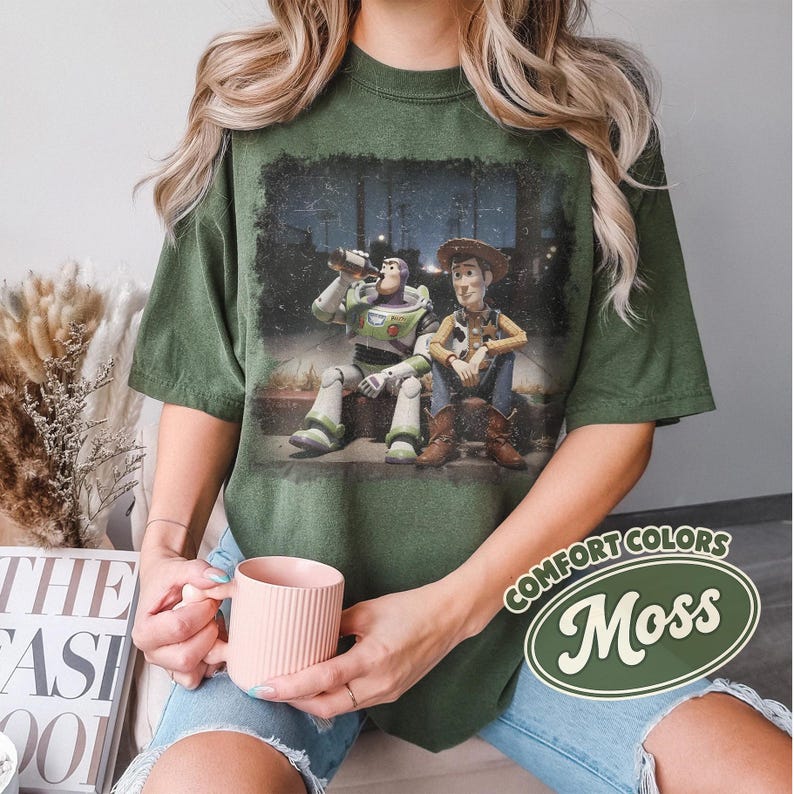 May include: Moss green t-shirt featuring a graphic of Buzz Lightyear and Woody from Toy Story. The characters are depicted sitting together. The shirt has a distressed look and the words "Comfort Colors" and "Moss" are printed on the shirt.
