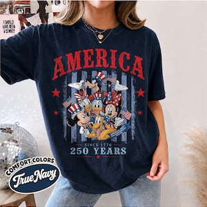 May include: A dark navy blue short-sleeve t-shirt featuring a graphic print of cartoon characters wearing patriotic hats. The text reads AMERICA, SINCE 1776, 250 YEARS, and AMERICA 250TH USA ANNIVERSARY. The design includes red stars and blue stripes.