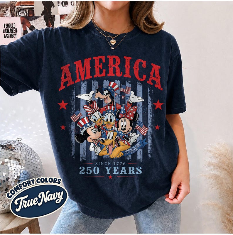 May include: A dark navy blue short-sleeve t-shirt featuring a graphic print of cartoon characters wearing patriotic hats. The text reads AMERICA, SINCE 1776, 250 YEARS, and AMERICA 250TH USA ANNIVERSARY. The design includes red stars and blue stripes.