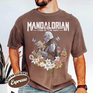 May include: Espresso-colored t-shirt featuring a graphic of The Mandalorian and Grogu surrounded by flowers and butterflies. The text "MANDALORIAN - THIS IS THE WAY" is printed across the top. The shirt has a vintage look.