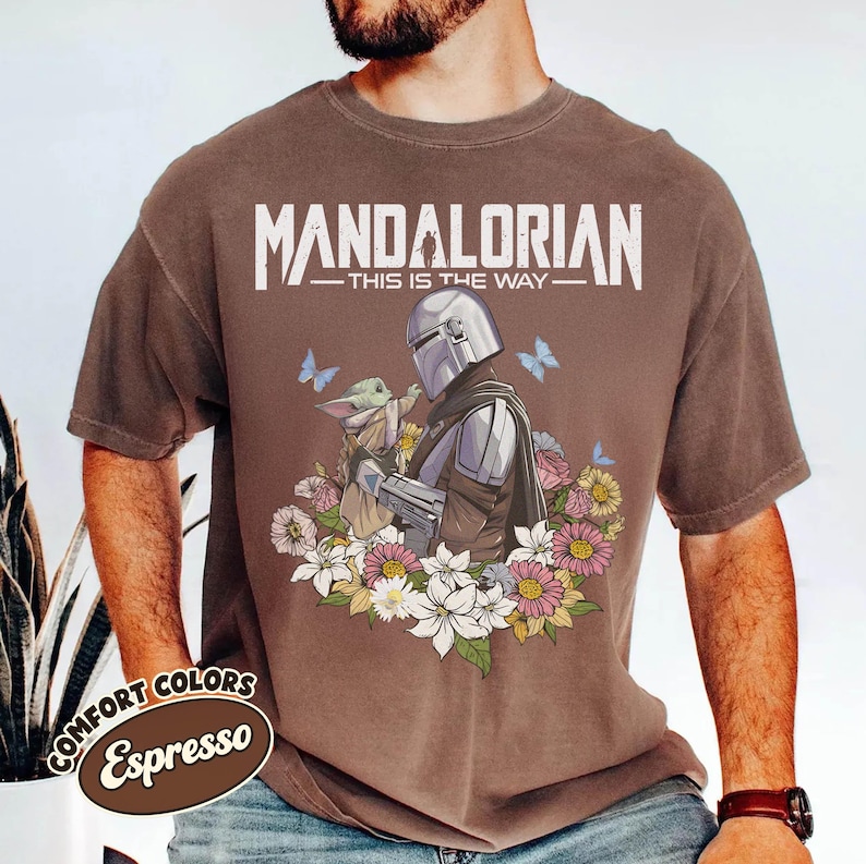 May include: Espresso-colored t-shirt featuring a graphic of The Mandalorian and Grogu surrounded by flowers and butterflies. The text "MANDALORIAN - THIS IS THE WAY" is printed across the top. The shirt has a vintage look.