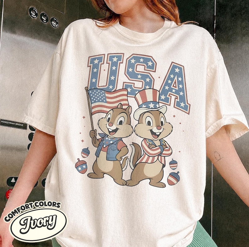 May include: Ivory-colored t-shirt featuring the word "USA" in blue with star accents. Two cartoon chipmunks are dressed in patriotic attire, one holding an American flag. Decorative red, white, and blue ornaments are scattered around the chipmunks.