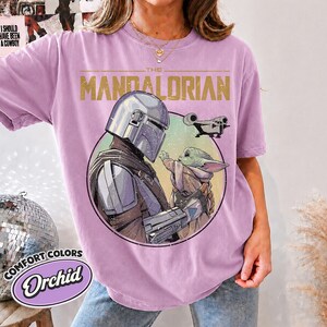 Puede incluir: Una camiseta de color orqu&iacute;dea con un estampado gr&aacute;fico de The Mandalorian y Grogu dentro de un marco circular. El texto THE MANDALORIAN est&aacute; impreso en dorado sobre los personajes, con una peque&ntilde;a ilustraci&oacute;n de una nave espacial.