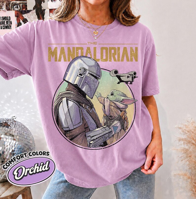 Puede incluir: Una camiseta de color orqu&iacute;dea con un estampado gr&aacute;fico de The Mandalorian y Grogu dentro de un marco circular. El texto THE MANDALORIAN est&aacute; impreso en dorado sobre los personajes, con una peque&ntilde;a ilustraci&oacute;n de una nave espacial.