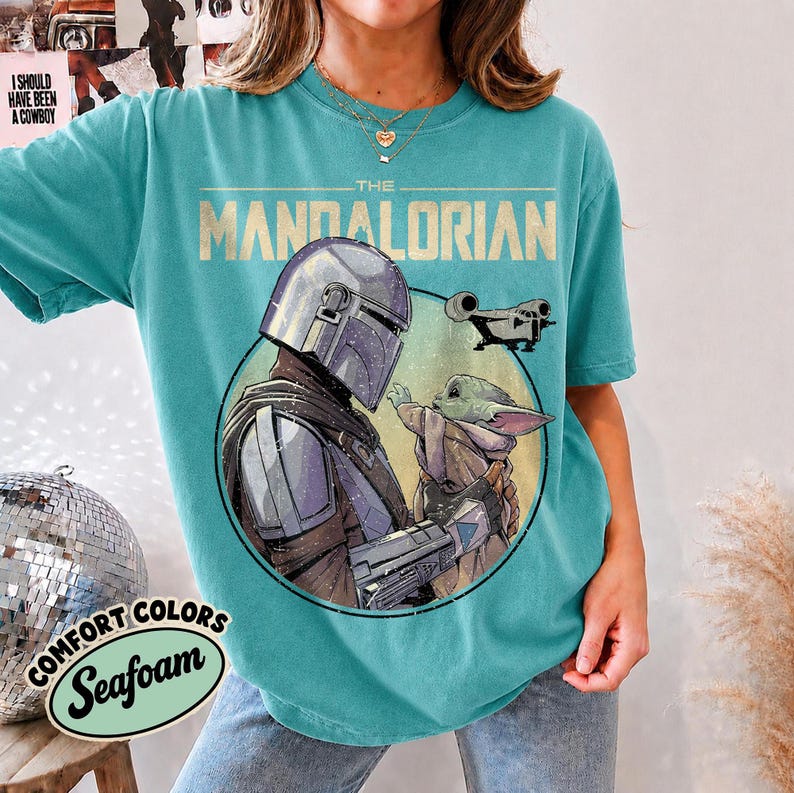 Puede incluir: Una camiseta de manga corta color verde azulado con un estampado gr&aacute;fico de The Mandalorian y Grogu dentro de un marco circular. El texto THE MANDALORIAN est&aacute; impreso en una fuente clara y llamativa sobre los personajes.